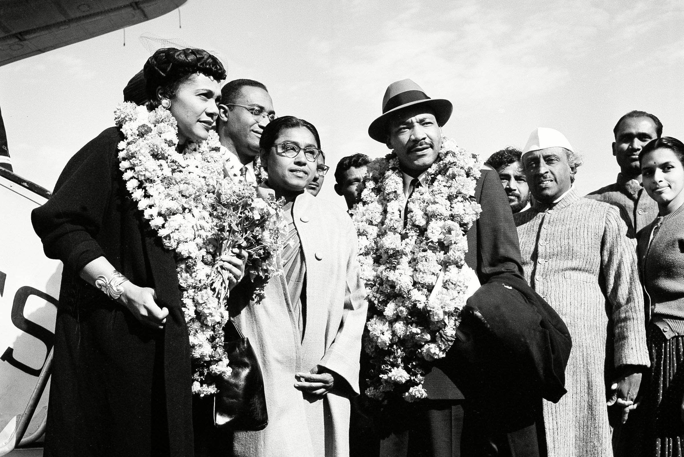 A group of people wearing flower garlands are gathered outdoors near an airplane, posing for a photo under a clear sky.