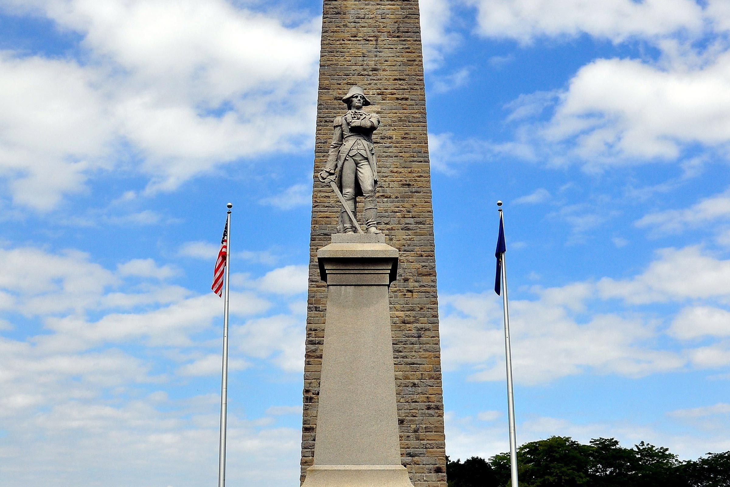 Stone statue stands on a monument pedestal, surrounded by two flagpoles, under a partly cloudy blue sky.