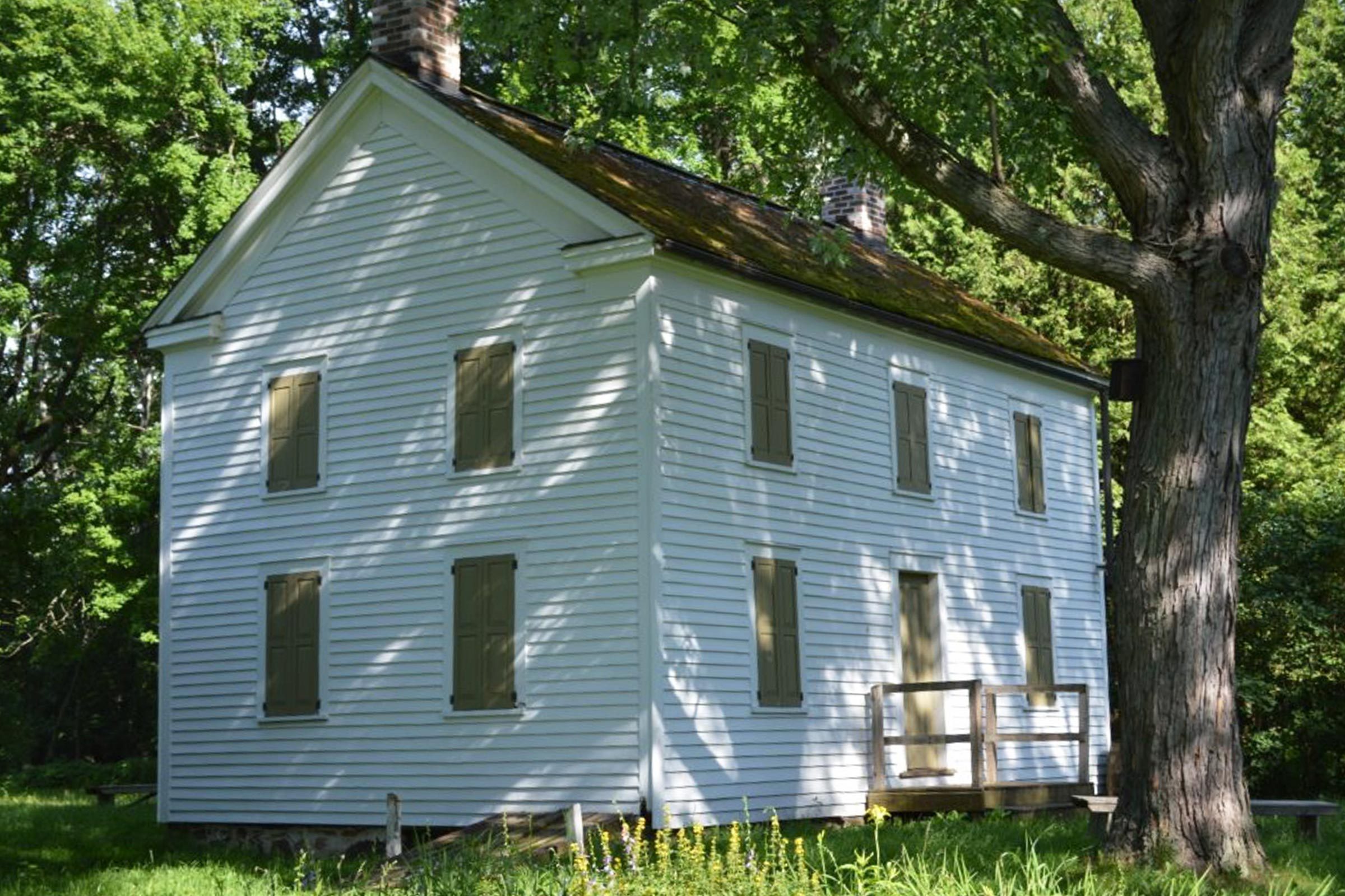 A white, two-story house stands quietly, surrounded by lush trees and greenery, casting shadows on the building's closed windows.