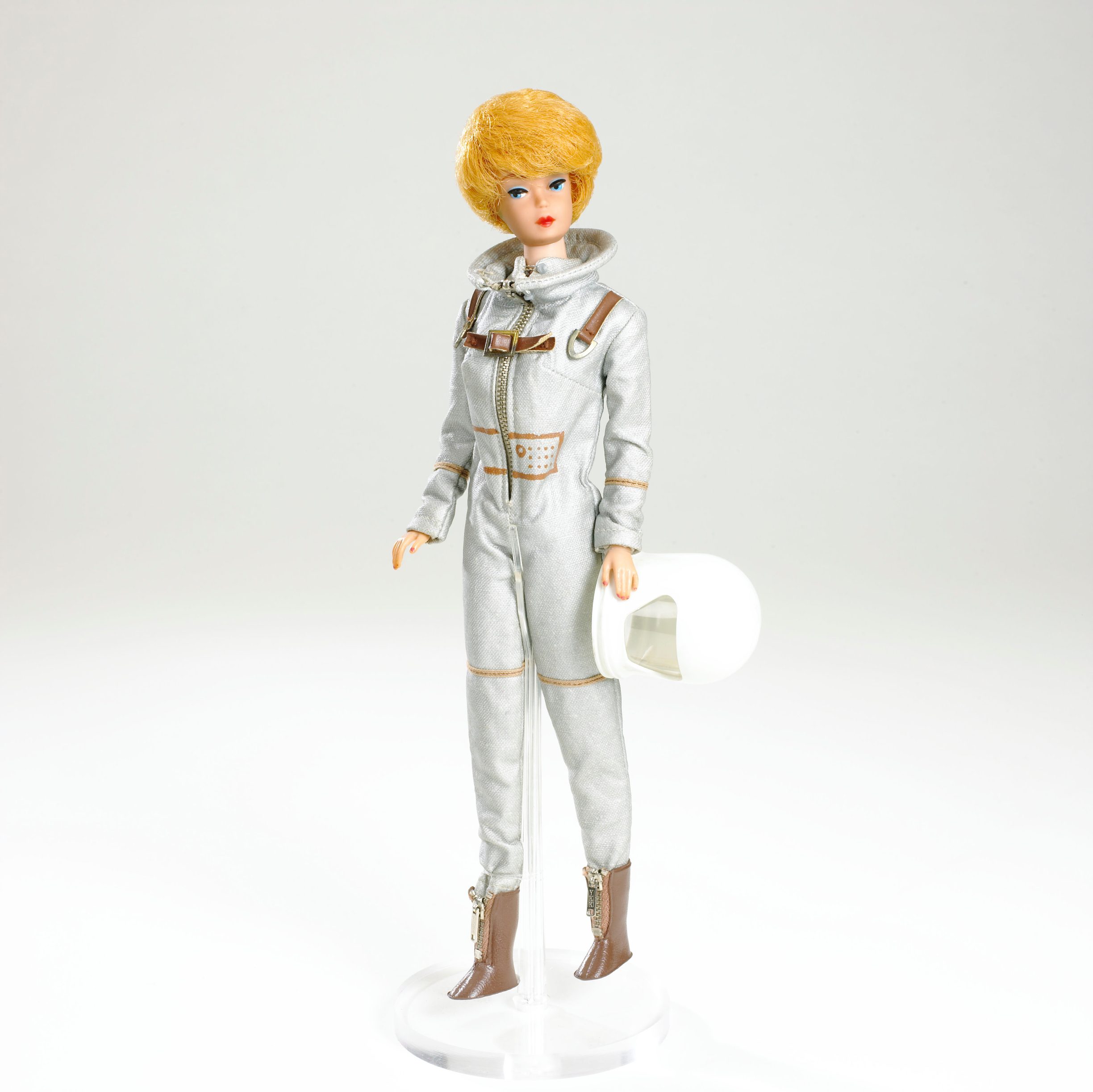 Doll in metallic space suit holds helmet, standing on a clear base against a plain white background.