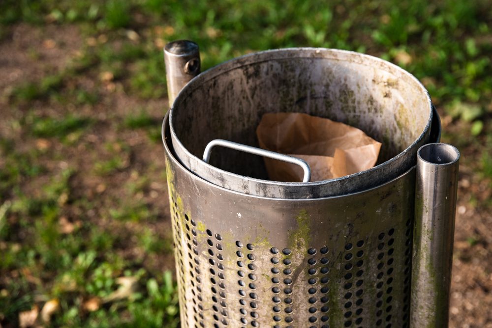 Metal trash can holds crumpled paper bag, situated in a park with grass and dirt path surrounding it.