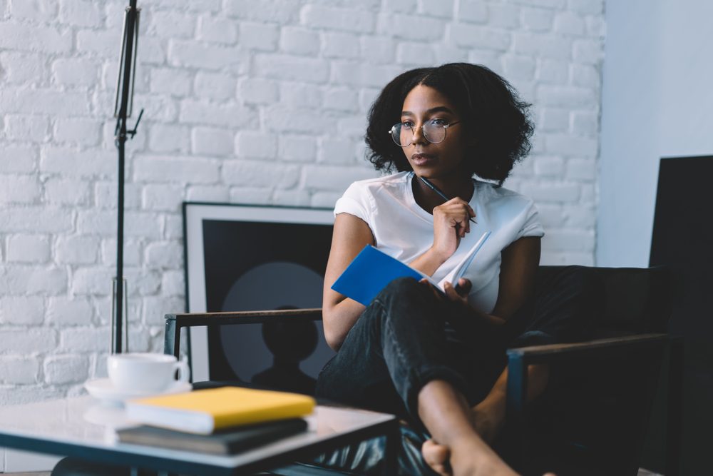 Thoughtful african american young woman in optical eyeglasses holding pen and thinking on creative ideas for writing her blog publication in notebook enjoying leisure time at home interior apartment