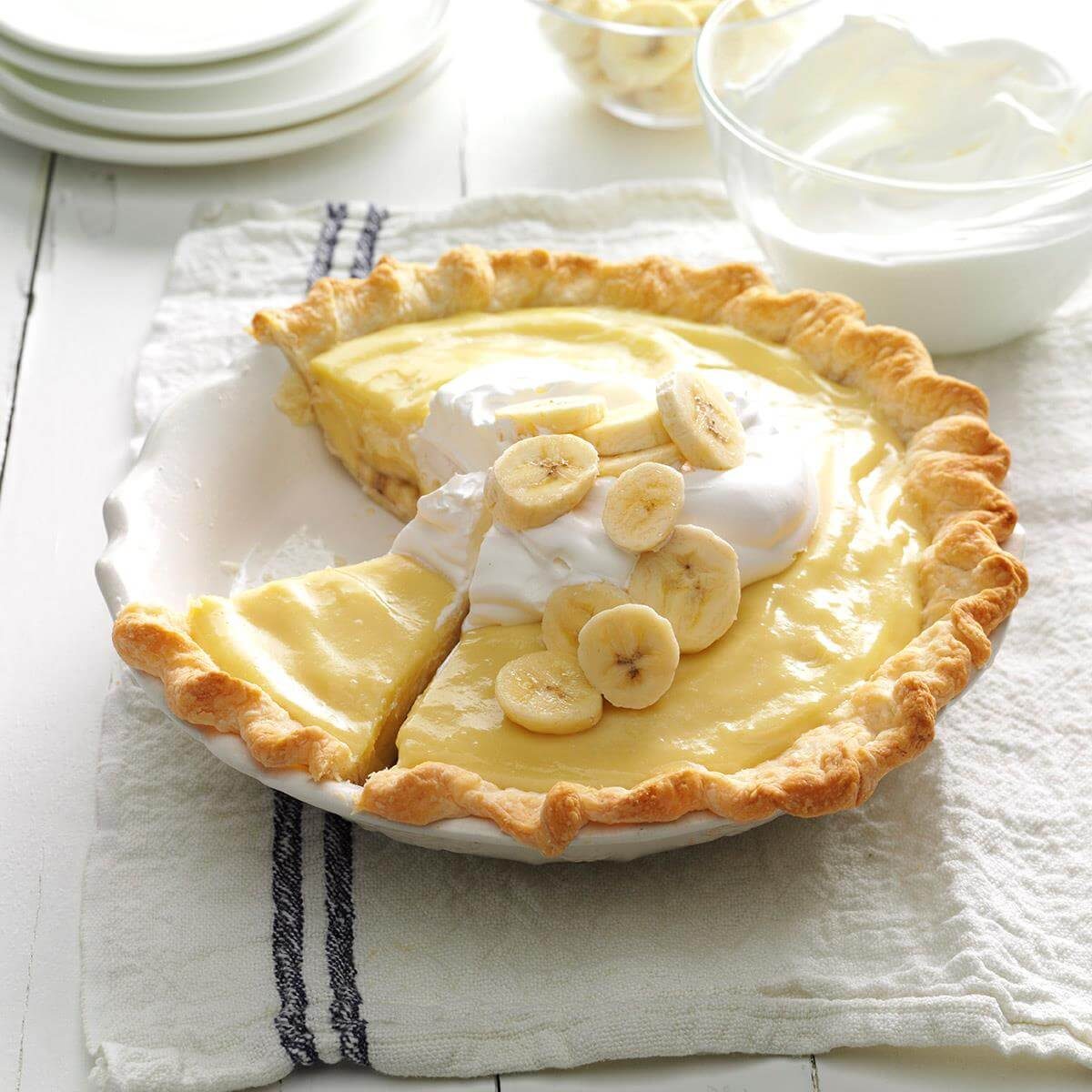 Pie topped with sliced bananas and whipped cream, served in a white dish on a textured cloth, with extra cream and plates nearby.