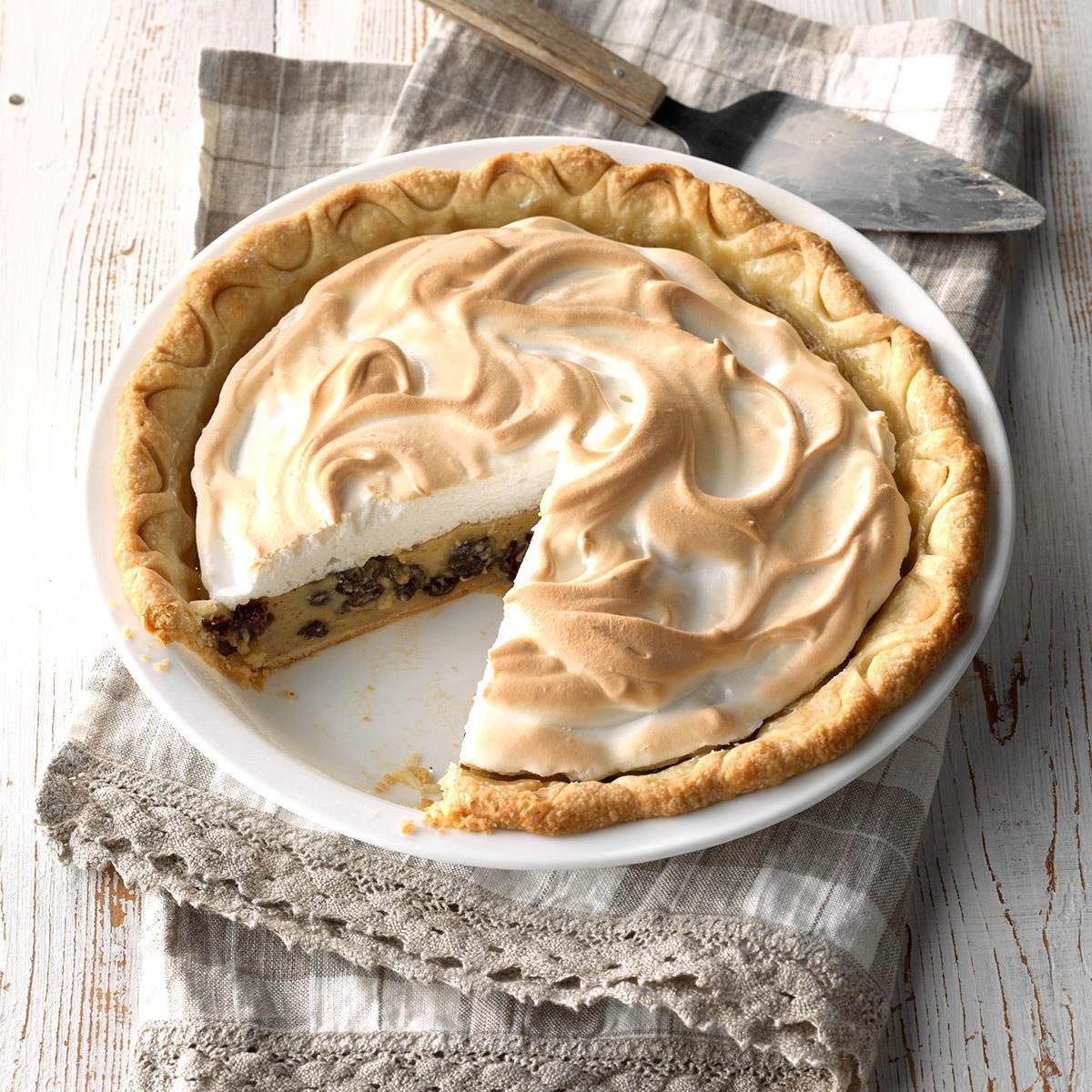 Pie sits with golden meringue, a slice missing, revealing a nutty filling, on a patterned cloth with a pie server nearby.