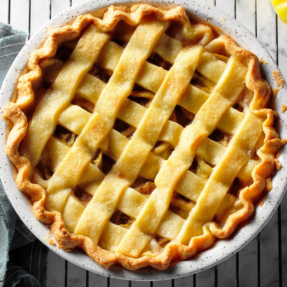 Pie with golden lattice crust cooling on wire rack, surrounded by a speckled pie dish and green cloth, suggesting a homemade setting.