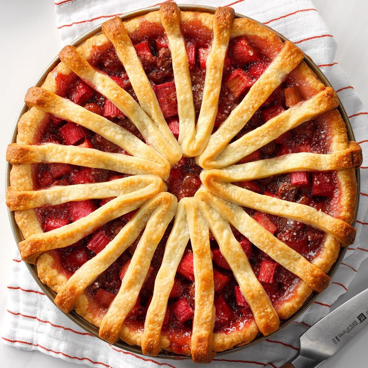 Pie with golden lattice crust radiates from center, filled with bright red fruit; set atop a striped cloth with a visible knife handle.