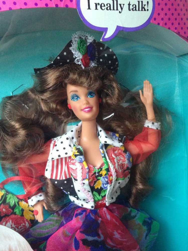 A fashion doll waves in vibrant floral attire, with polka-dot accents inside a colorful box. Text reads, "I really talk!"