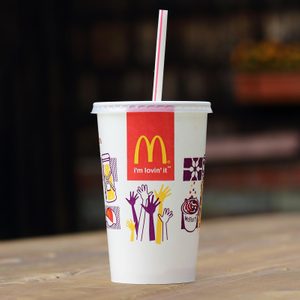 A McDonald's cup with a straw sits on a wooden table outdoors, featuring graphics and text: 
