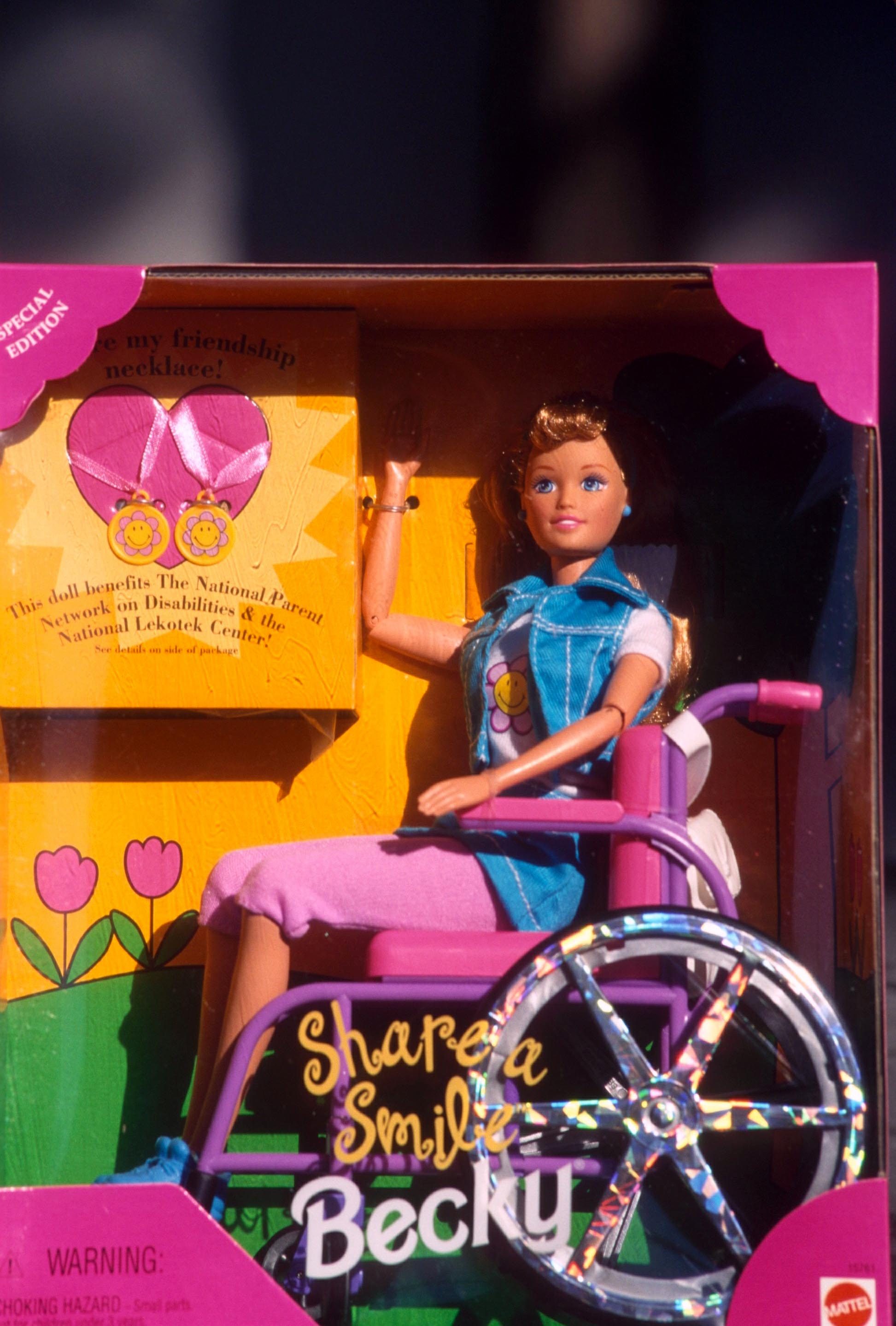 A doll in a wheelchair smiles in packaging, wearing casual clothes. Text: "Share a Smile Becky" and indicates support for disabilities organizations.