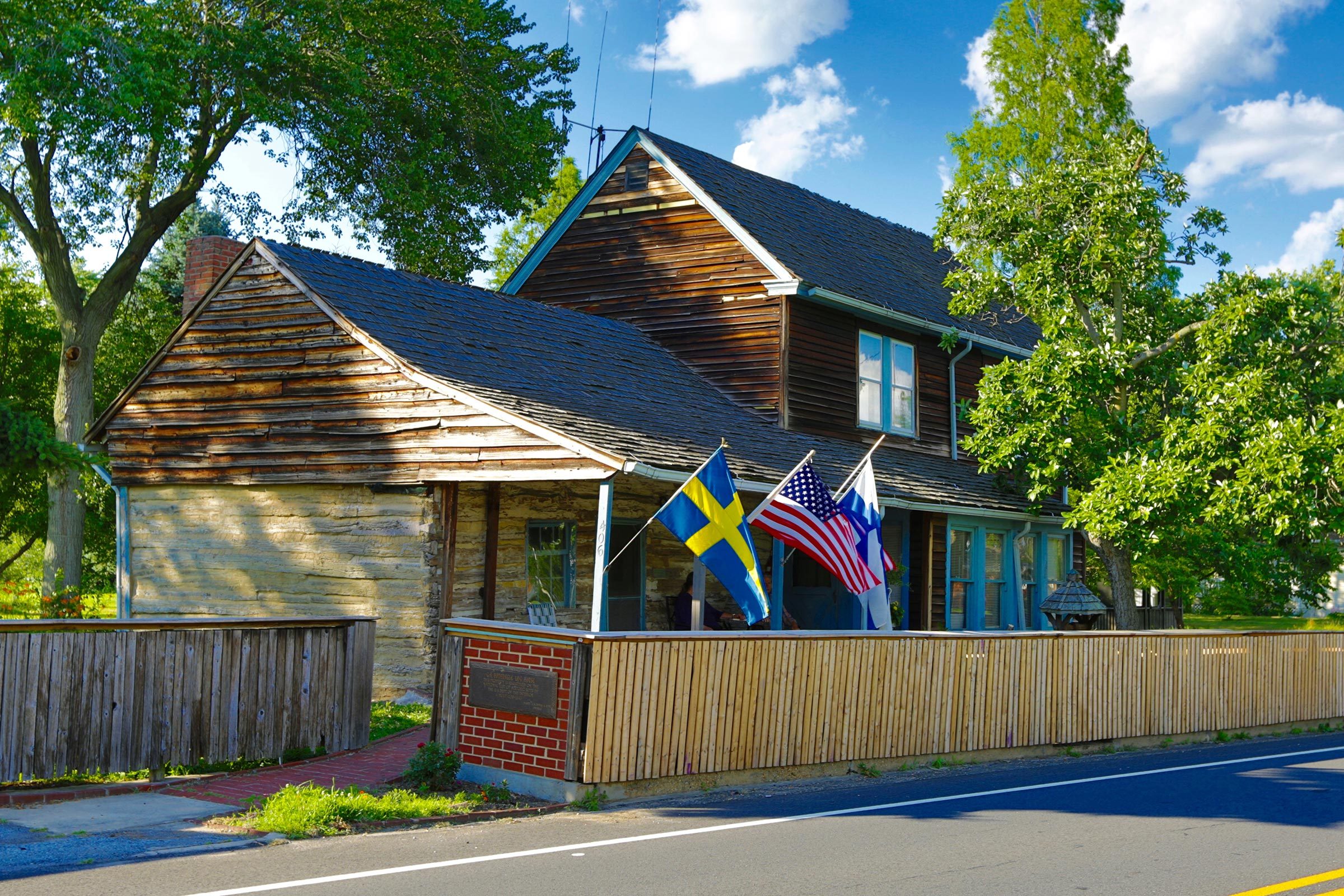 Wooden house displays Swedish, American, and Finnish flags on porch, with a rustic fence, surrounded by lush trees under a blue sky with clouds.
