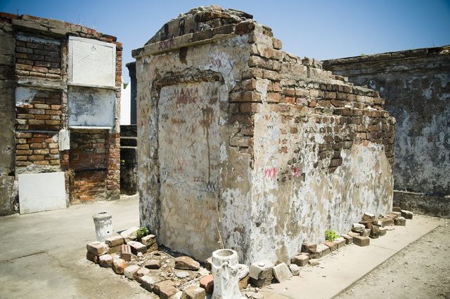 Weathered brick structure displaying graffiti, standing in an expansive, sunlit cemetery with scattered stone fragments on the ground.