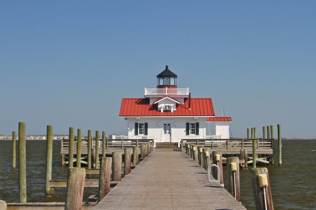 Lighthouse sits at pier's end, surrounded by water, under a clear blue sky, with red roof and white walls.