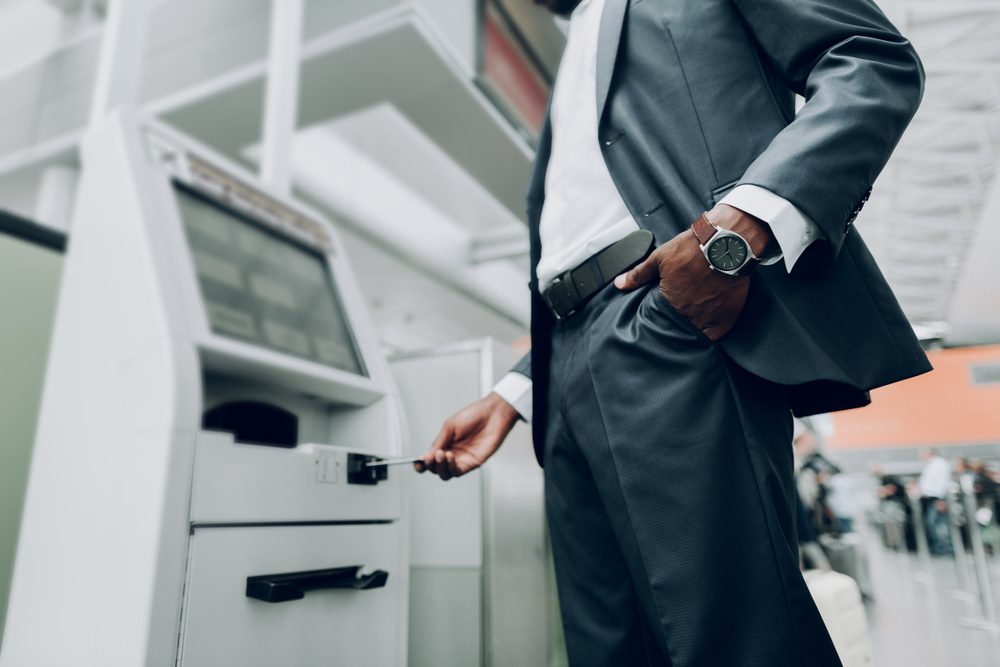 Person inserts card into machine at a modern airport terminal, dressed in a suit with hand in pocket.