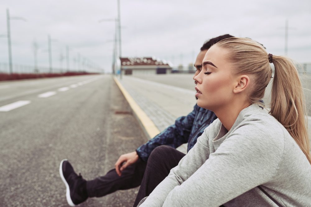 Two people sit relaxed on a deserted road, with a cloudy sky and distant buildings in the background.