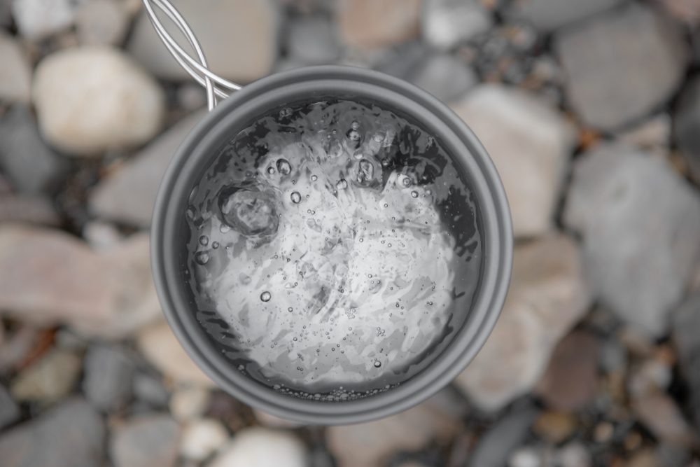 Metal Gray Mug With Boiling Water Close-Up On Background Of River Stones, Top View. Boiling Water In The Campaign.