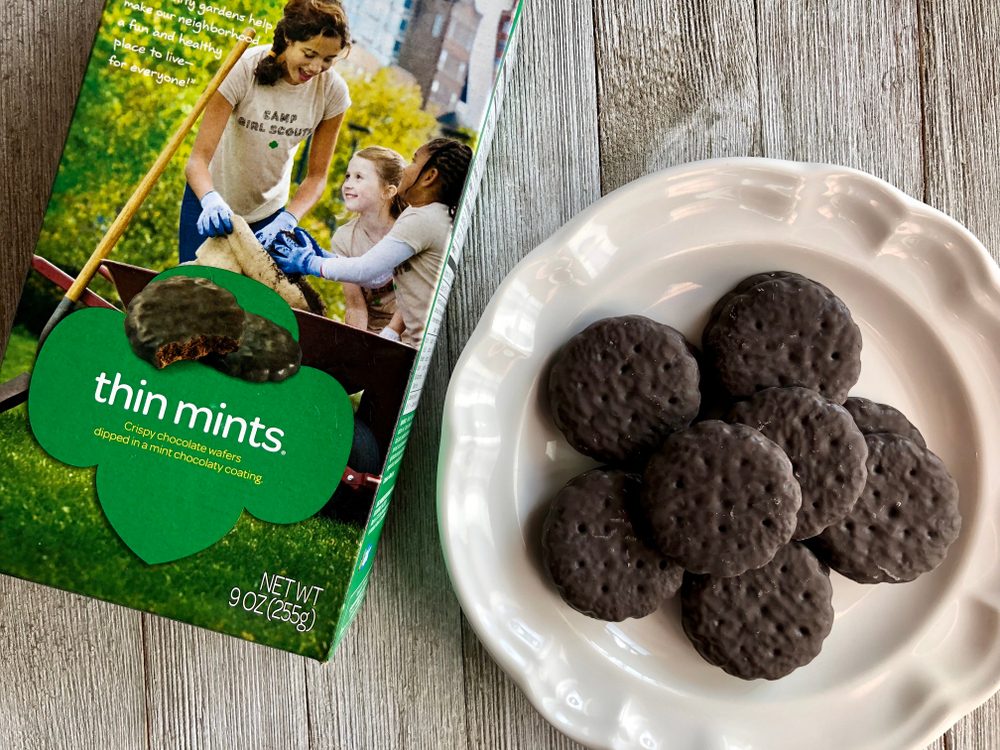 Cookies rest on a white plate beside a box with a girl gardening, emphasizing "Thin Mints" in green text.