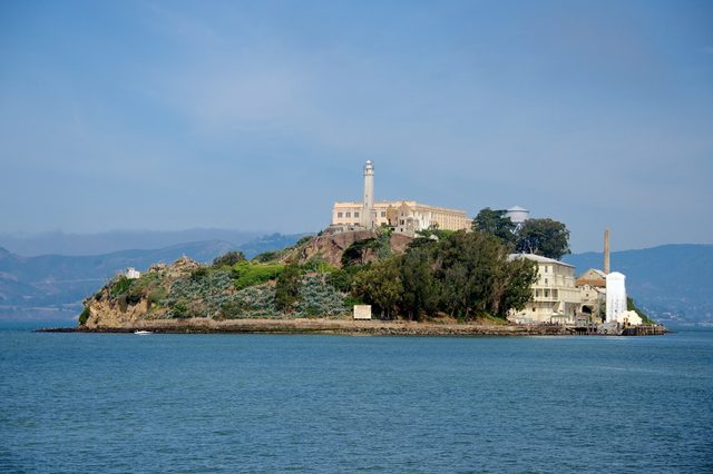Island with a large building and lighthouse sits quietly, surrounded by water, with mountains visible in the distant background under a clear sky.