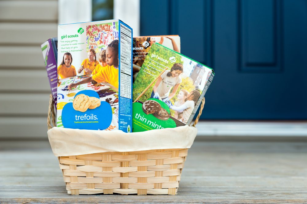 Basket holds Girl Scouts cookie boxes, featuring Trefoils and Thin Mints, placed on a wooden porch in front of a closed blue door.