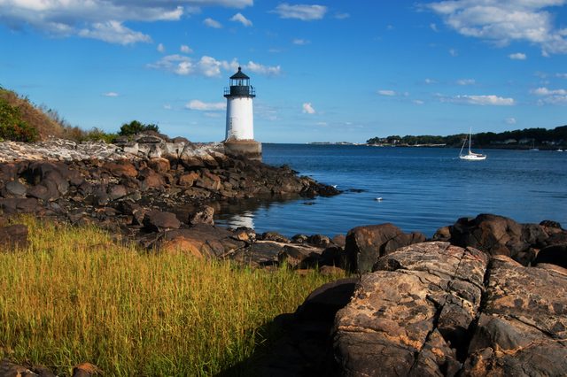 Lighthouse stands still on rocky shore, overlooking calm sea with sailboat in the distance, under a clear blue sky with scattered clouds.