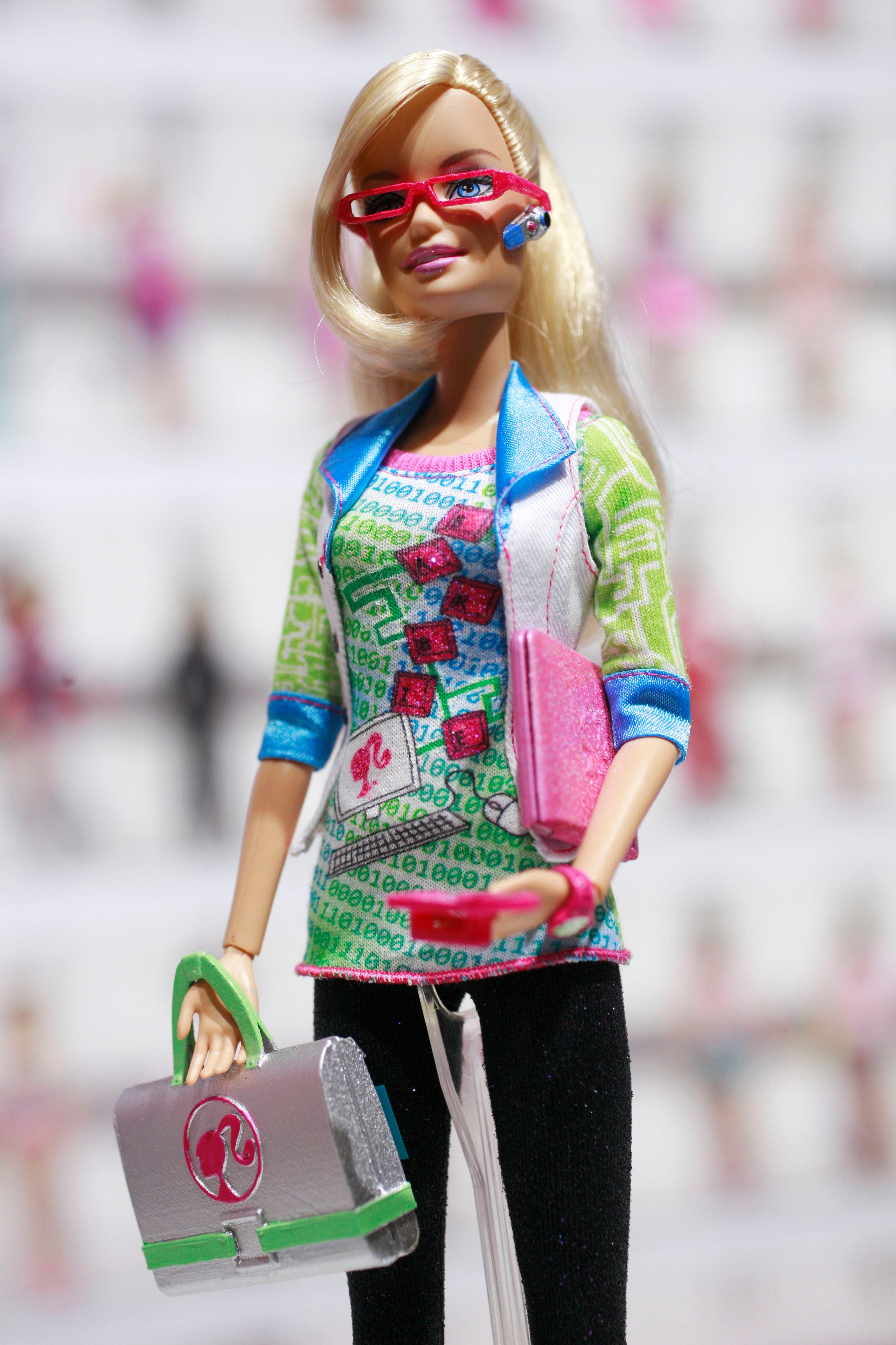 Doll wearing colorful tech-themed attire and glasses, stands holding a pink device and silver laptop bag in a bright, blurry background.