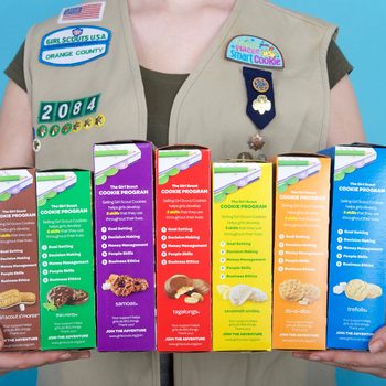 Boxes of Girl Scout cookies are held by a person in a Girl Scout vest against a blue background. Text lists cookie names and program skills.