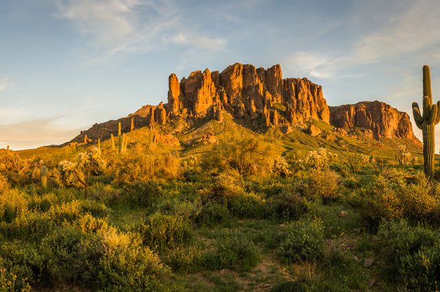 Rock formation sits illuminated by golden sunlight, surrounded by desert vegetation and tall cacti, under a partly cloudy blue sky.