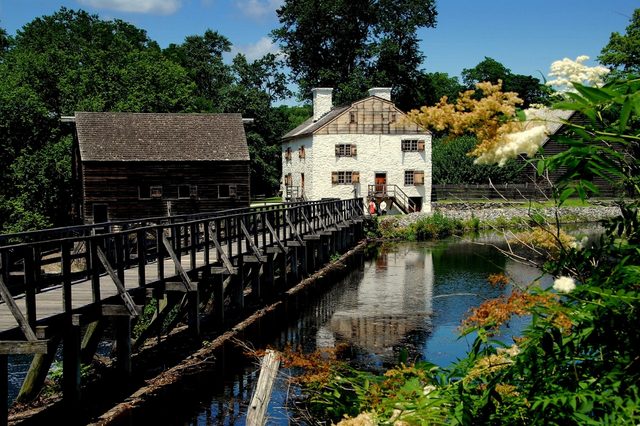 A wooden bridge crosses a reflective pond, leading to rustic buildings surrounded by lush greenery and flowers under a clear blue sky.