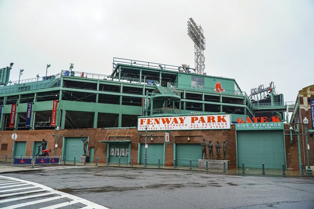 Stadium entrance displays "FENWAY PARK" sign, surrounded by statues and gates, under a cloudy sky, with nearby street visible.