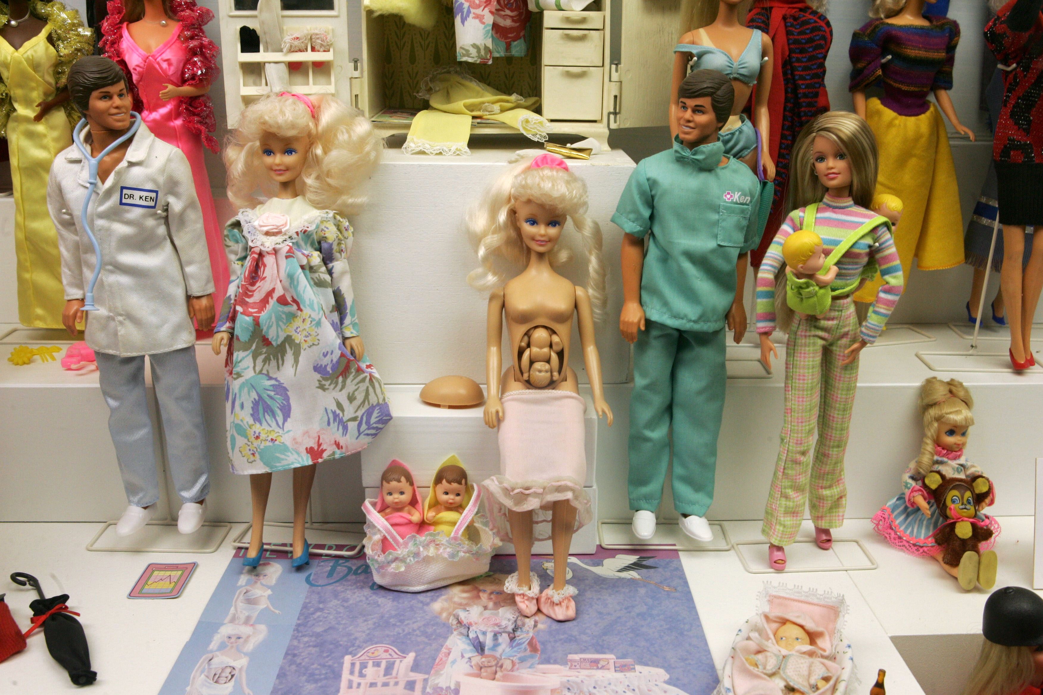 Dolls stand on display, featuring a pregnant doll with removable belly; surrounded by medical-themed dolls and baby accessories.
