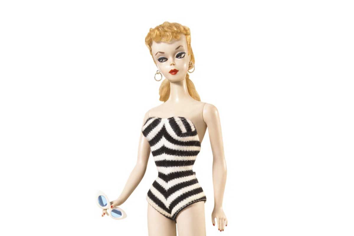 A doll wears a black-and-white striped swimsuit, holding sunglasses, with gold hoop earrings, against a plain white background.