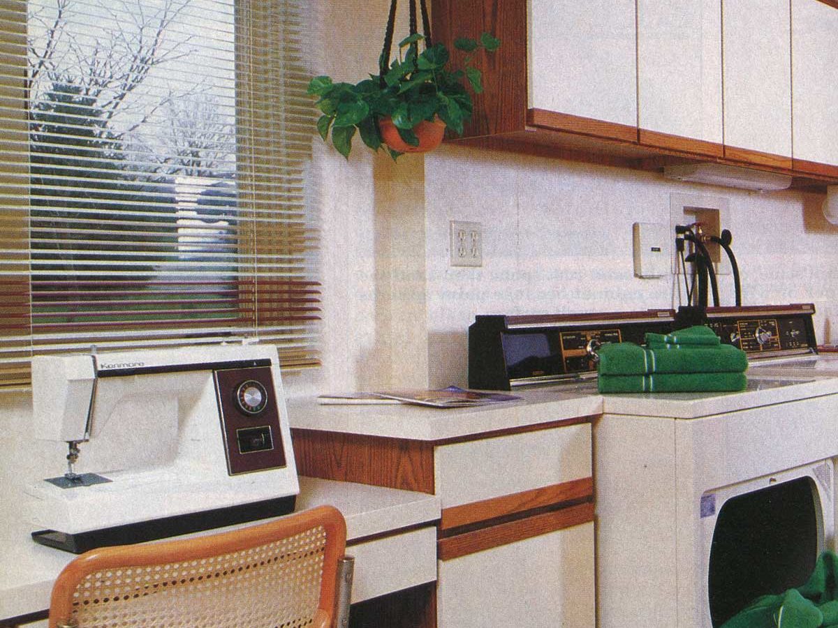 Sewing machine sits on a counter beside a washing machine in a well-organized laundry room with a hanging plant and window blinds.