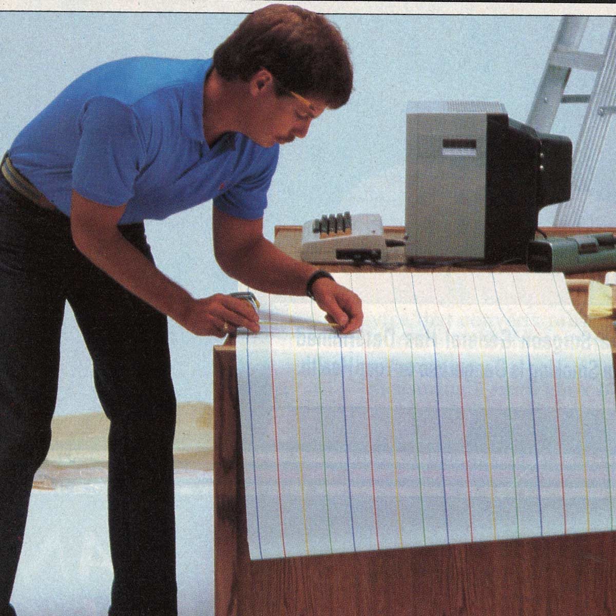 A person reviews long, colored graph paper on a desk with a calculator and printer in a bright room.