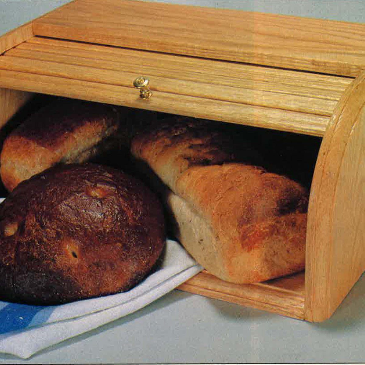 A wooden bread box with a roll-top lid holds various loaves, surrounded by a kitchen countertop setting.