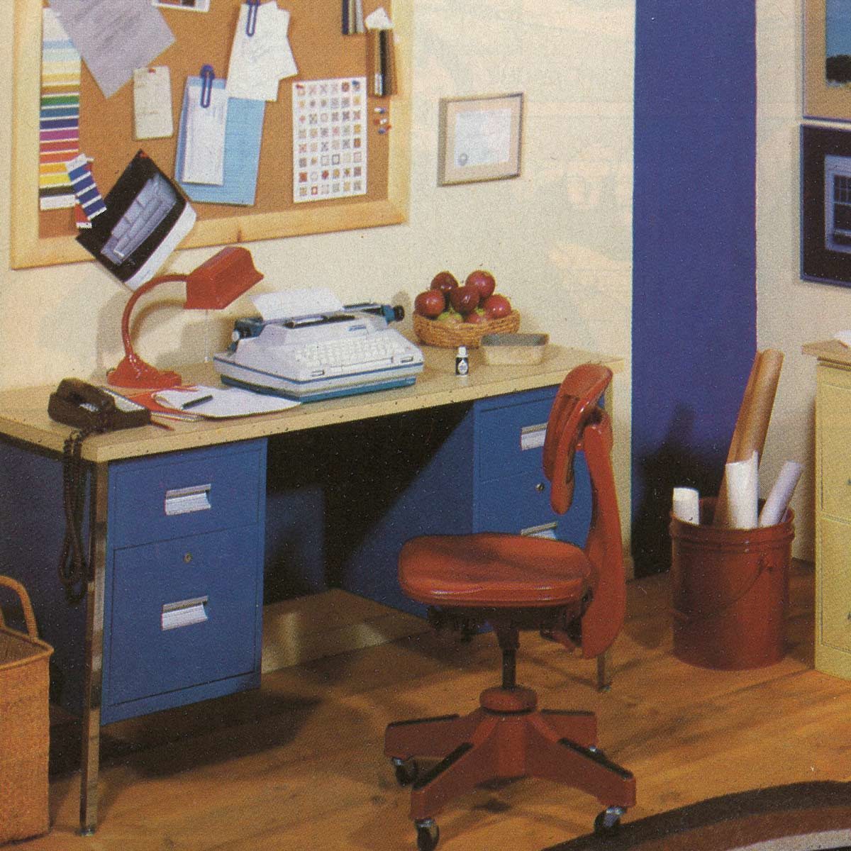 Chair sits in front of a desk holding a typewriter, red lamp, and basket of apples in a neatly arranged office space.