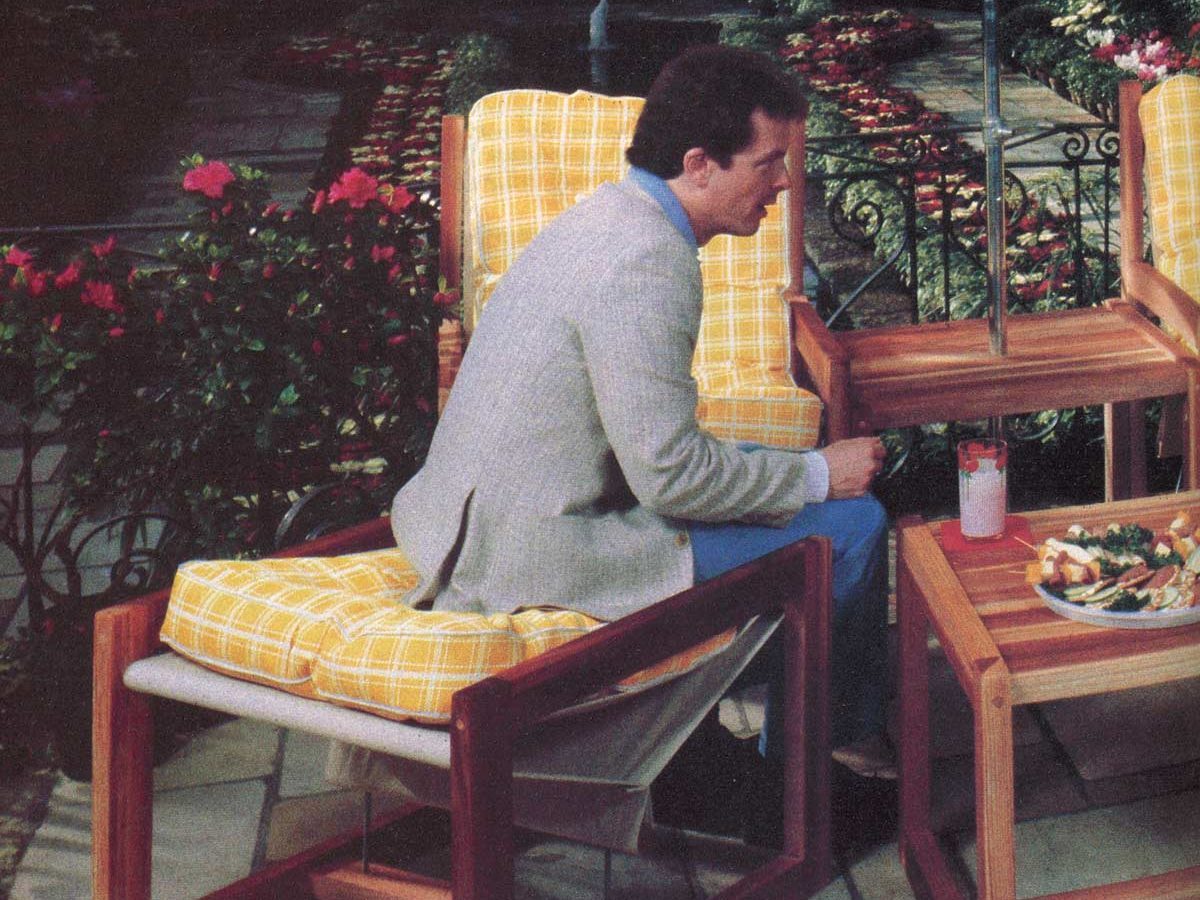 Man sits on cushioned chair, drinking at wooden table, surrounded by vibrant garden and checkered umbrella overhead.