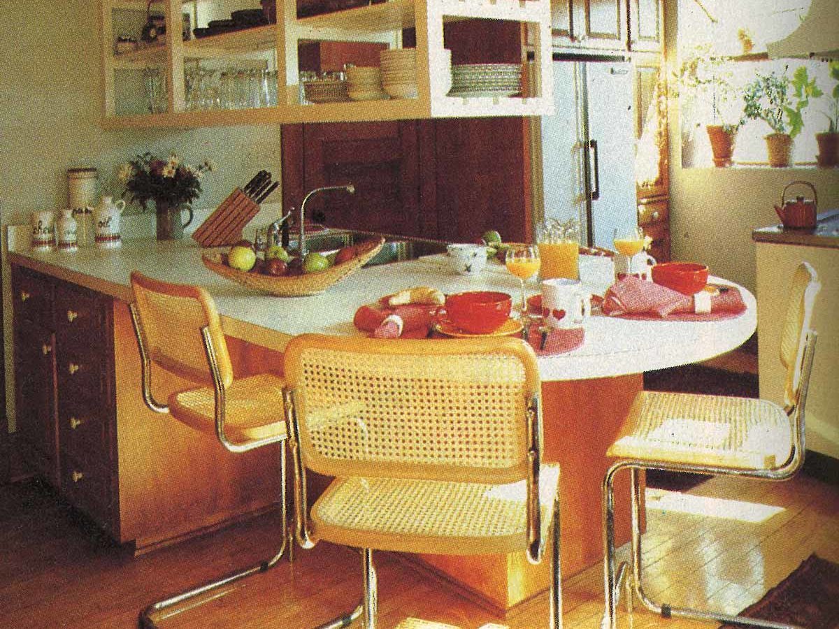 Kitchen table set for breakfast, featuring chairs and dishes with food, in a sunlit room with open shelves and potted plants.