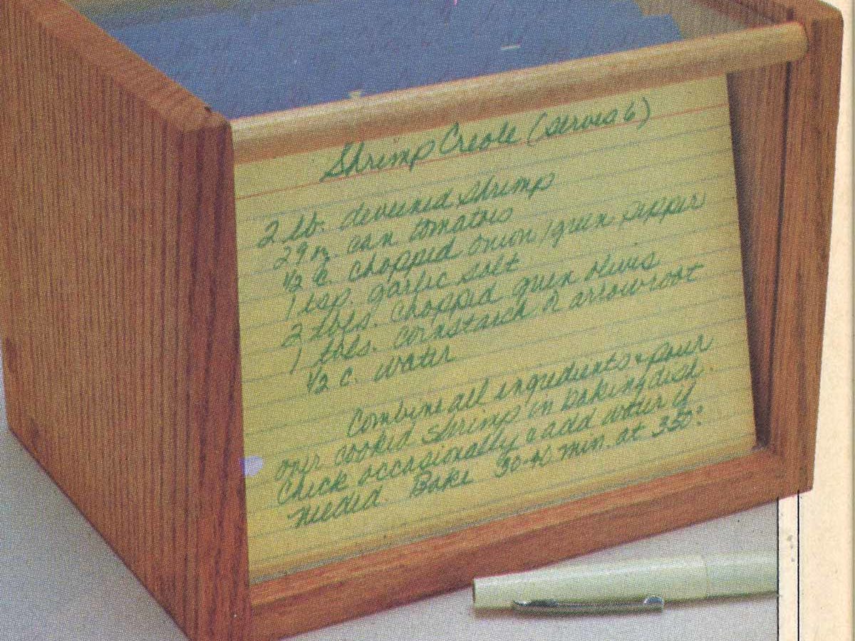 Wooden box holds handwritten recipe cards, arranged upright. A pen rests nearby on a table, with text detailing a Shrimp Creole recipe.