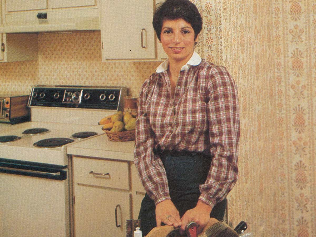 Woman stands holding toolbox, smiling, in a vintage kitchen with floral wallpaper and white cabinets.