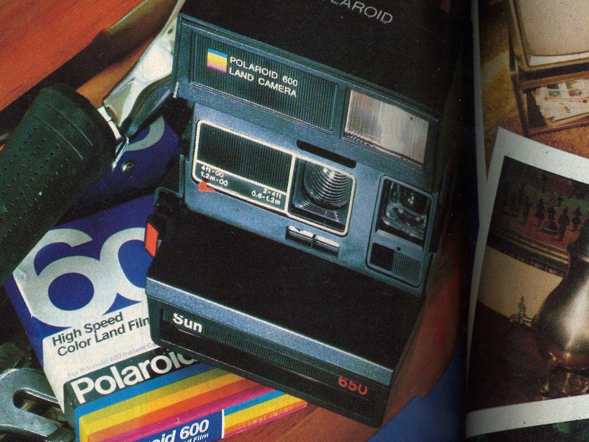 Polaroid camera rests on a wooden surface with colorful film packages nearby, alongside photos and a flashlight.