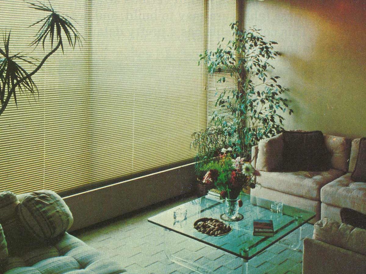 Glass table holds vase of flowers, surrounded by beige sofas and plants, in a softly lit room with large window blinds and carpeted floor.