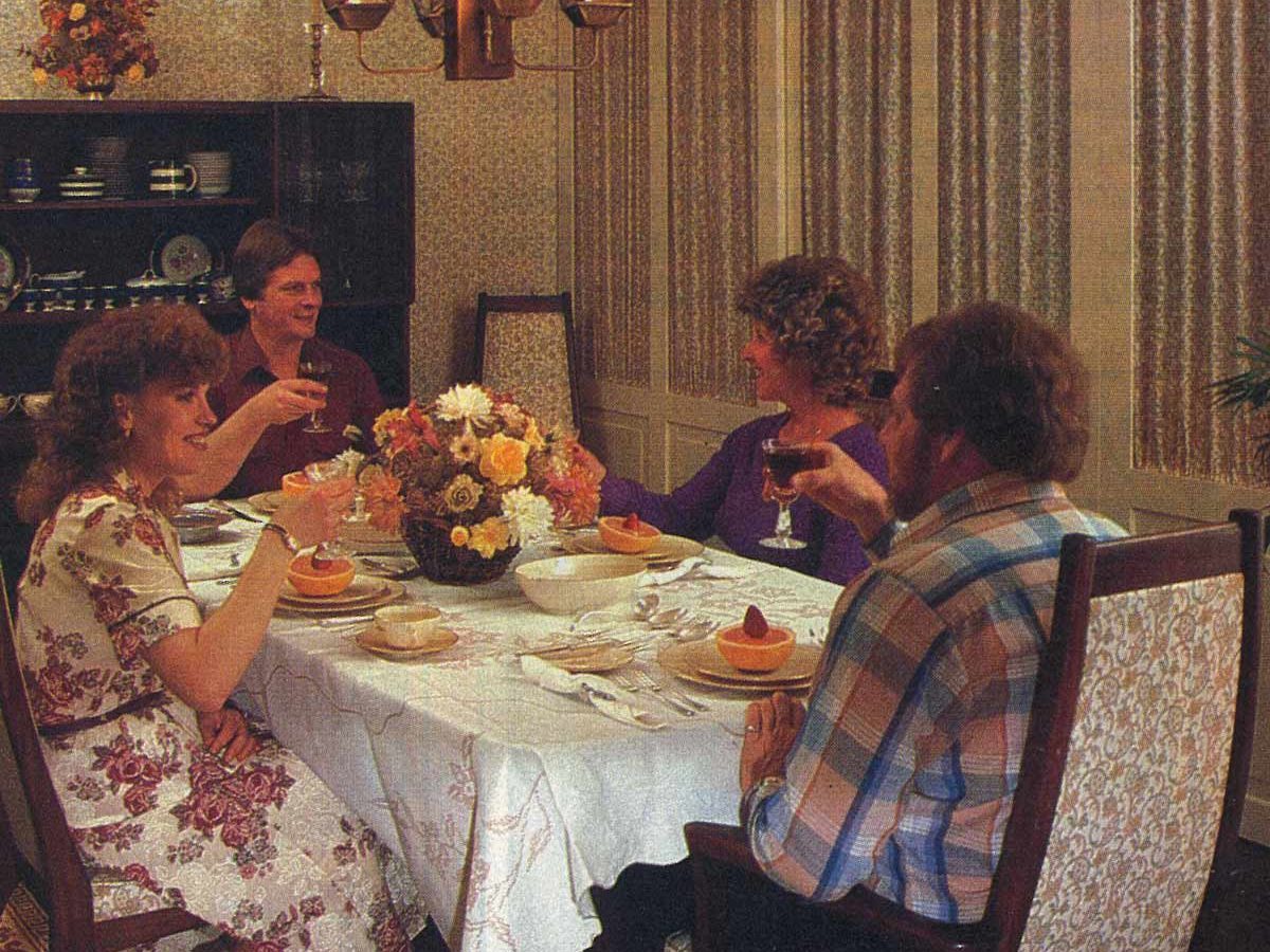 People sit dining; they cheerfully hold drinks around a flower-adorned table in a warmly lit, elegant dining room with patterned walls and chandelier.