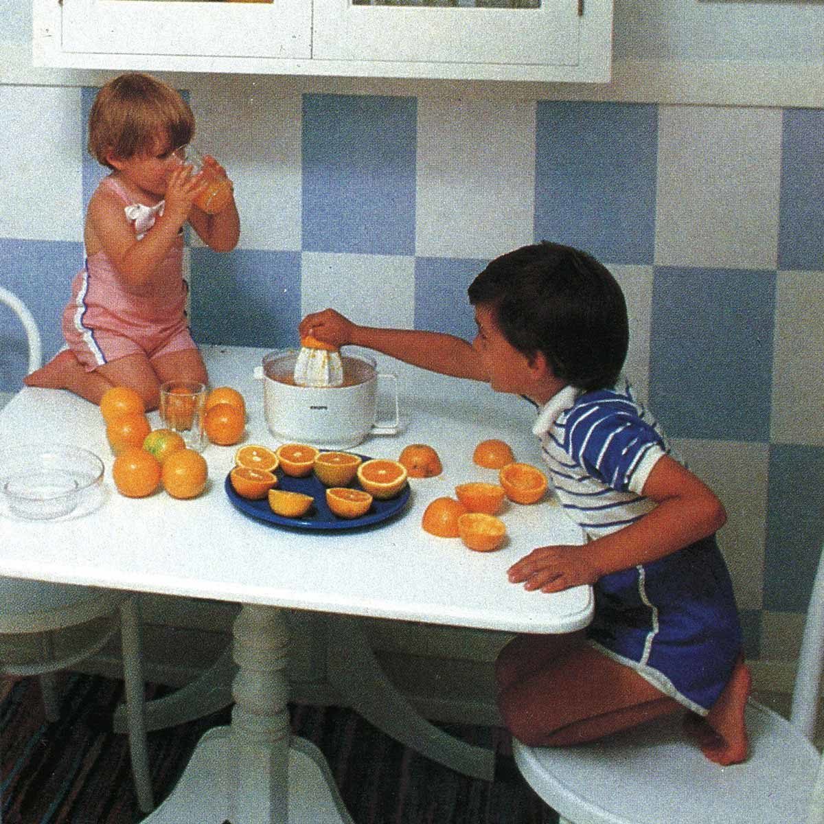Children make orange juice at a white table, surrounded by halved oranges, in a kitchen with blue checkered walls.