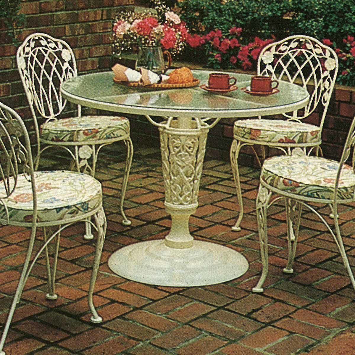 Round table with pastries and coffee, surrounded by ornate chairs, set on a brick patio with blooming flowers in the background.