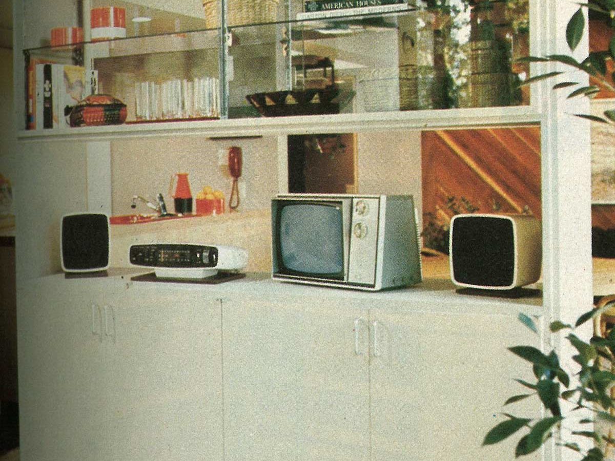 A vintage television sits on a white cabinet, flanked by speakers; kitchen items and books are on shelves above, with plants nearby.