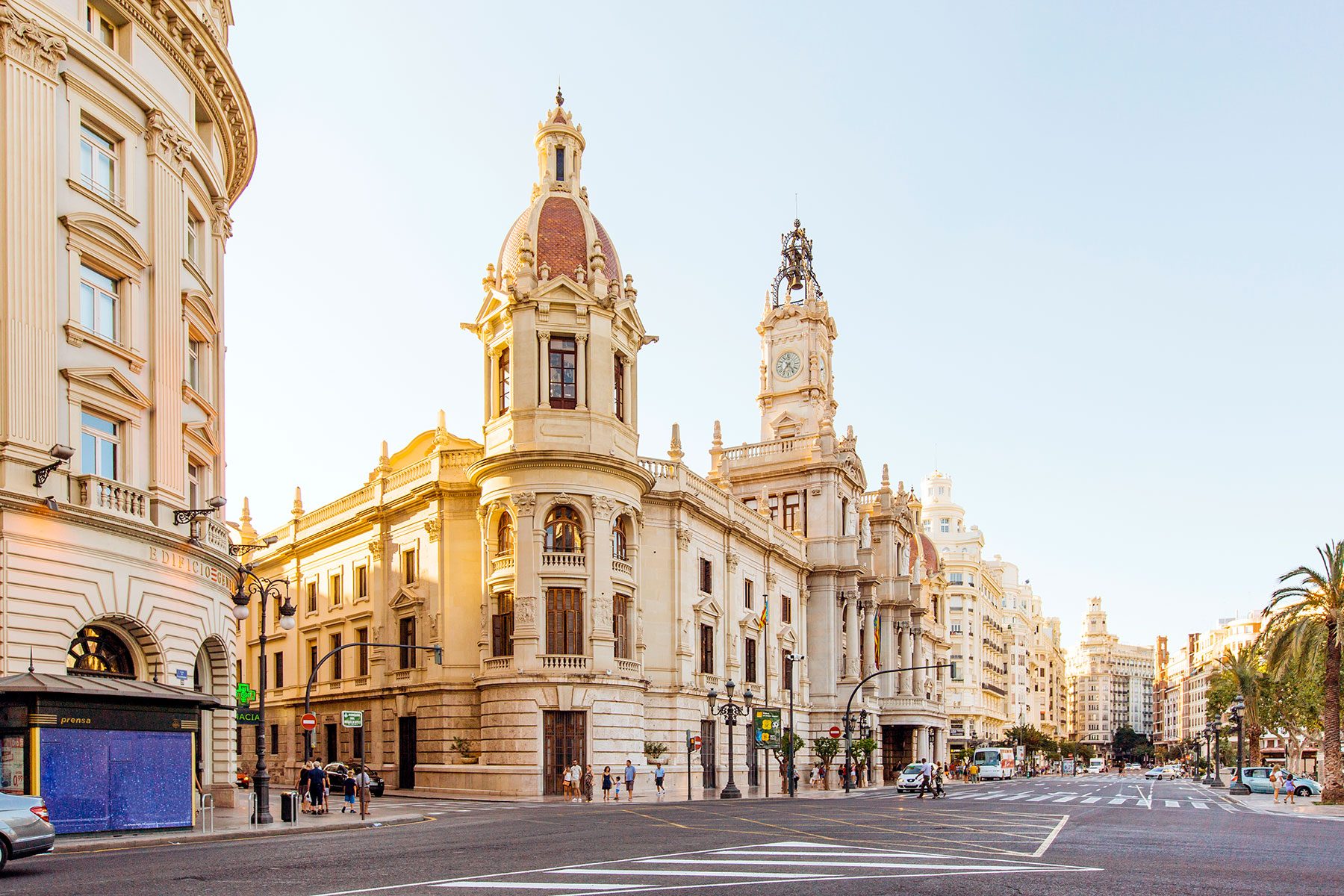 Historic buildings with domes and clock tower loom over a bustling city street lined with shops and lampposts under a clear blue sky.