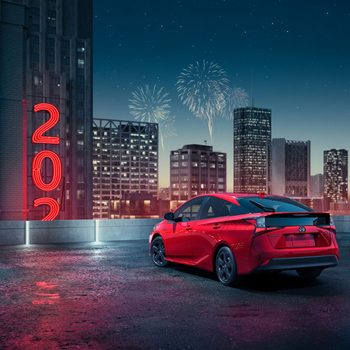 A red car parked on a rooftop, surrounded by city skyscrapers and fireworks. Bright red neon numbers "2023" glow against a night sky.