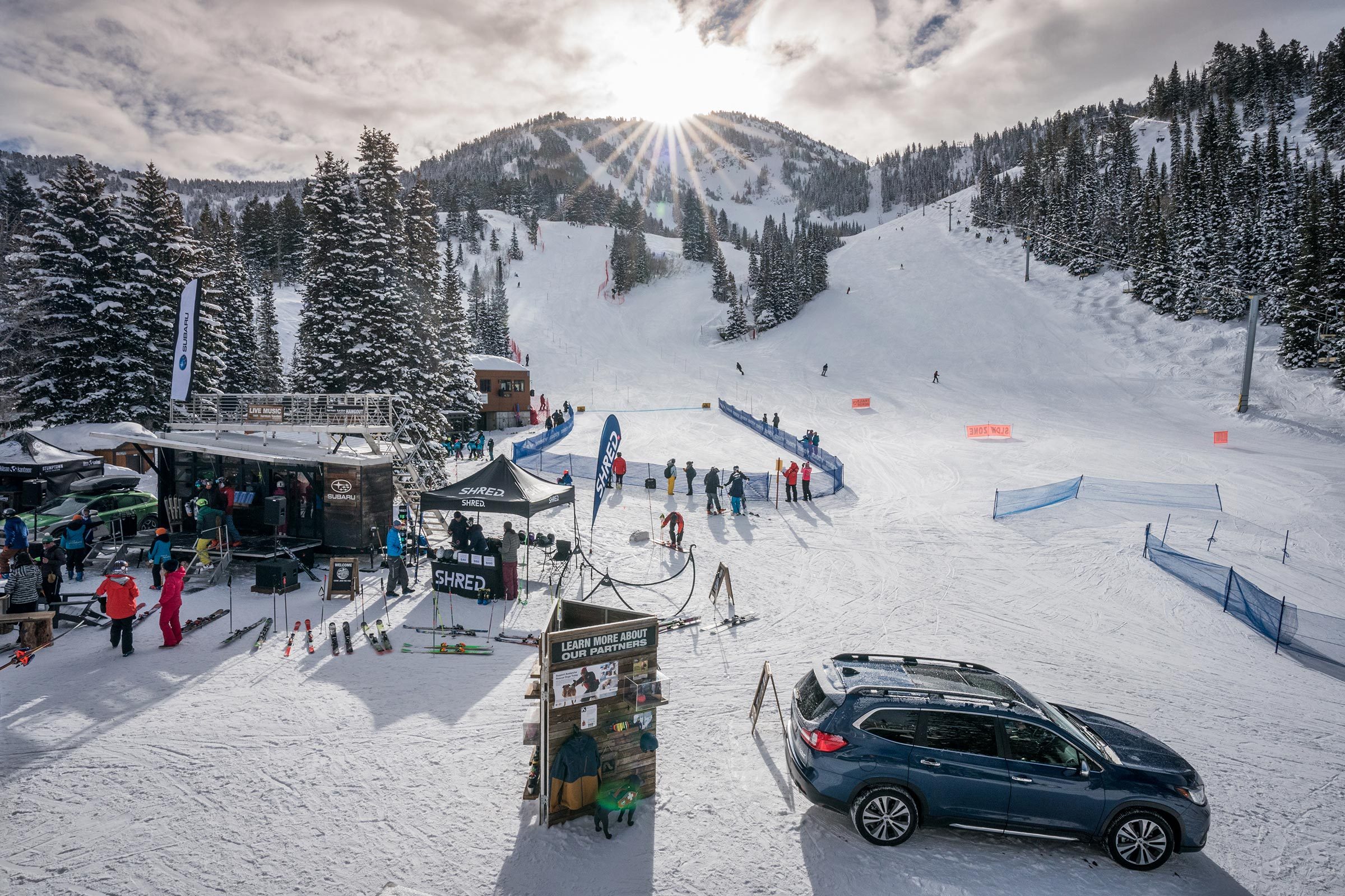 Skiers descend a snow-covered slope surrounded by pine trees, with event tents and parked car nearby, under a sunlit, cloudy sky.