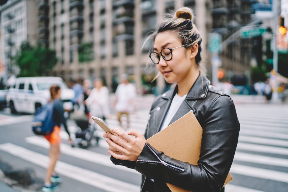Person reading phone while holding a folder, standing in a busy city crosswalk with blurred pedestrians and buildings in the background.