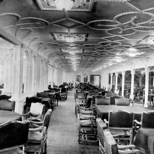 Chairs and tables are neatly arranged in a lavishly decorated dining room with ornate ceiling patterns, surrounded by columns and large windows.