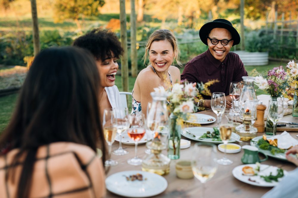 People are laughing and enjoying a meal at a table set for a garden party, surrounded by wine glasses and colorful flowers.